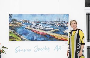 Emma Jacobs Artist standing next to one of her seaside paintings with signage Emma Jacobs Art